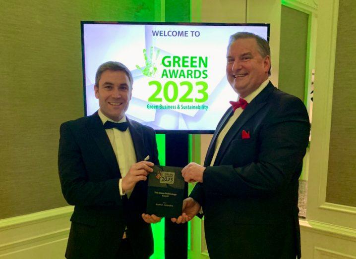 Two men in suits holding an award with a TV screen behind them, with "Green Awards 2023" visible on the screen.