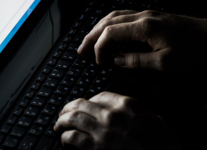 A pair of hands typing on a keyboard in a dark location, with the light from a computer screen visible on the left.