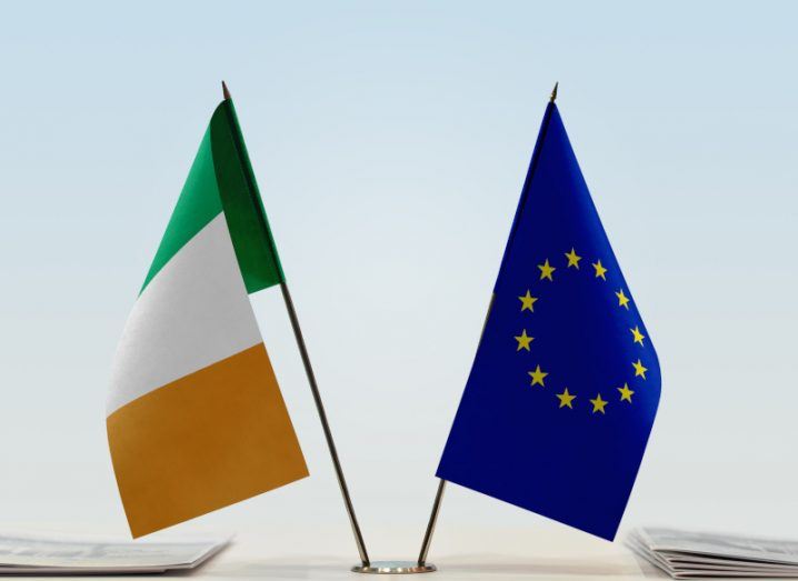 A flag of Ireland and a flag of the EU, next to each other on flag poles connected to a building.