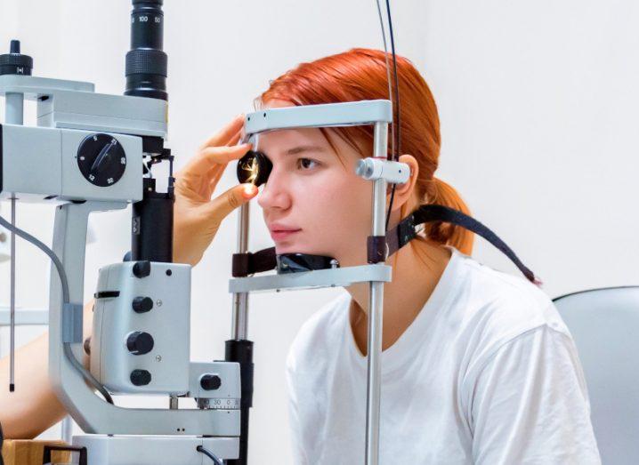 A woman with red hair sitting in front of an eye examination device, while another person's hand operates the device.