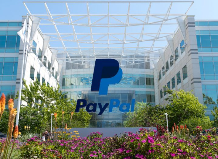 The PayPal logo in front of a large building, with plants surrounding the PayPal sign and a blue sky in the background.
