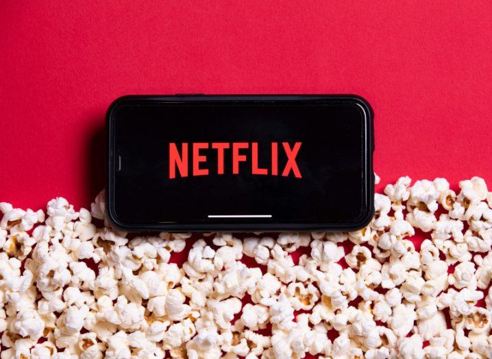 The Netflix logo on a smartphone screen, laying on a red background with a pile of popcorn below it.