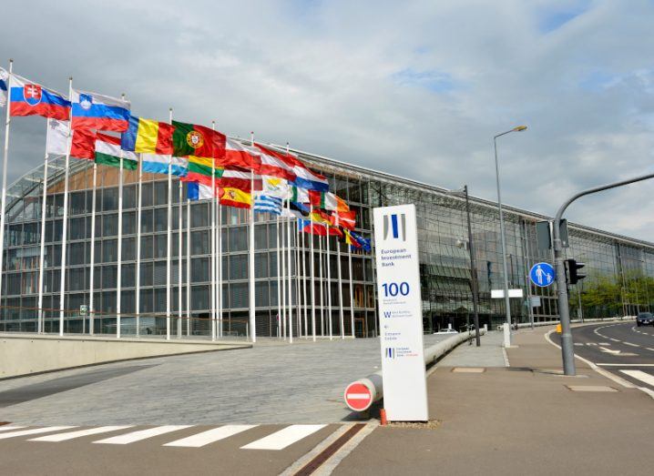 The European Investment Bank (EIB) building, with multiple country flags on flag poles in front of it and a cloudy sky above.