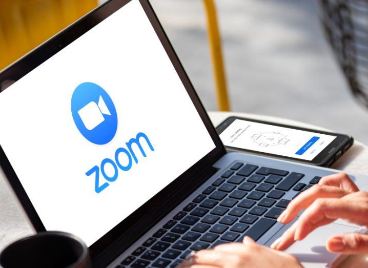 The Zoom logo on a laptop screen, with a person typing on its keyboard. A phone and cup are on either side of the laptop.