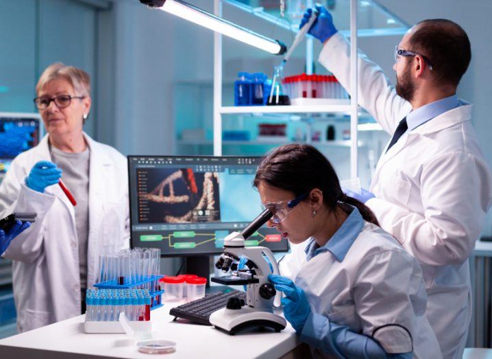 A group of scientists in a medical lab working together. One researcher is looking through a microscope while another is putting liquid into a vial. An image of a DNA strand is on a laptop screen on a table.