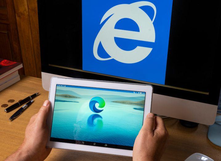 A person holding a tablet with the Microsoft Edge logo on the screen. A computer screen with the Internet Explorer logo is behind the tablet, laying on a wooden table.