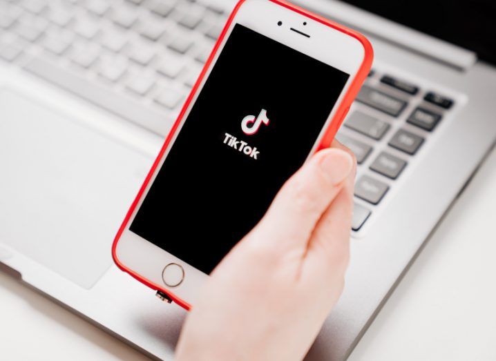 The TikTok app logo on a mobile phone screen, being held in a person's hand with a laptop under the phone.