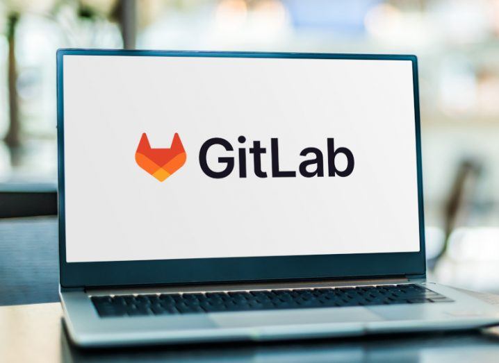 The GitLab logo on a laptop screen. The laptop is on a reflective table with a blurred interior in the background.