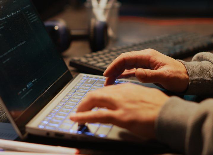 A person typing on a laptop keyboard that is on a wooden table, with another keyboard in the background.