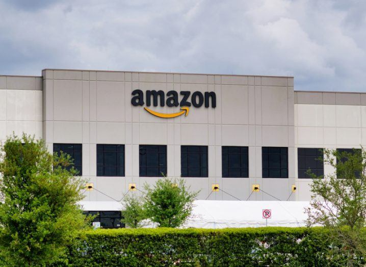 Amazon logo near the top of a large grey building, with green trees in the foreground and a cloudy grey sky in the distance.