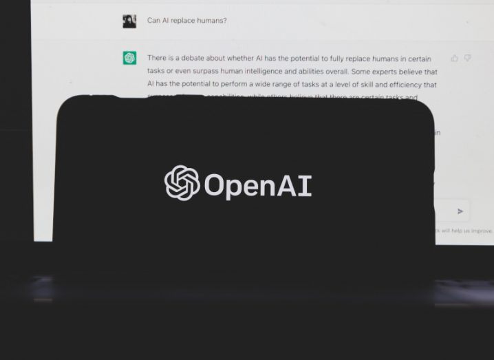 The OpenAI logo on a smartphone screen, with a computer screen behind it showing the ChatGPT chatbot.