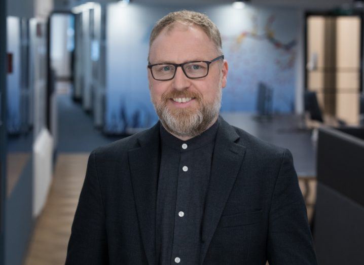 A man stands in the hallway of what appears to be an office, smiling at the camera. He is Ciaran McGuinness, a client partner at NTT Data UK&I.