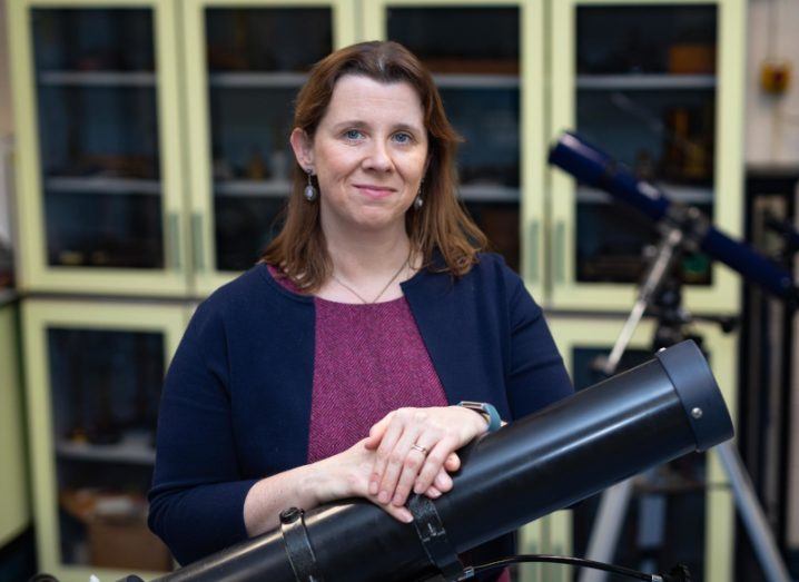 A woman with dark hair stands in front of a telescope smiling at the camera. She is astrophysicist Emma Whelan.