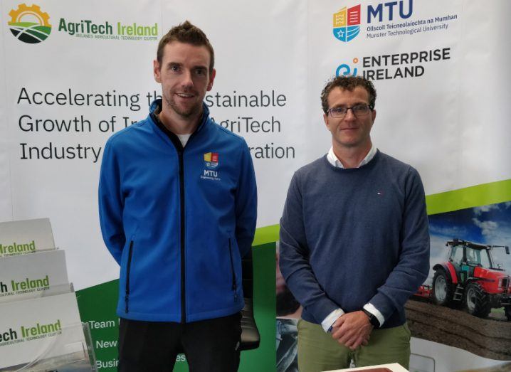 Two men standing next to each other, with signs in the background with the MTU and AgriTech Ireland logos on them.