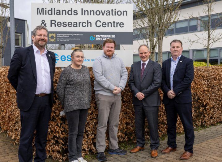 Five people standing outside a building with Midlands Innovation Research Centre written on a sign outside it. Autumn leaves in the background on a hedge behind the people.