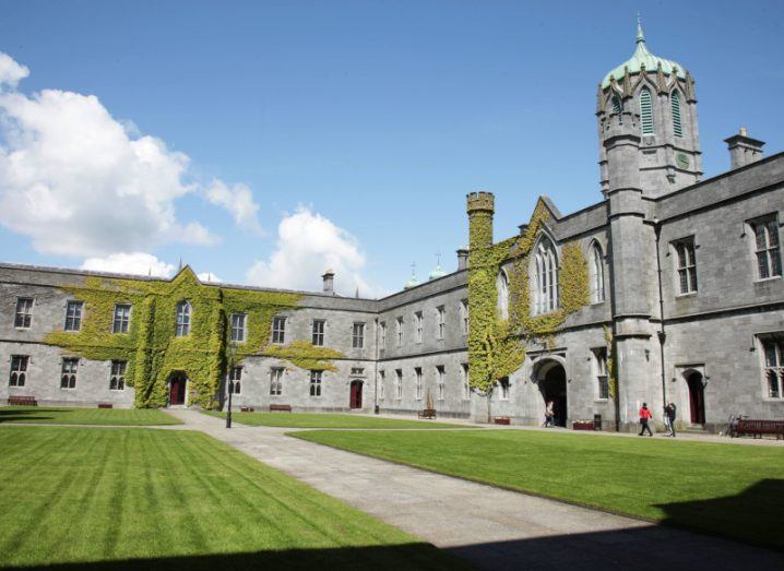 The Quadrangle building of University of Galway, with a grey footpath surrounded by grass in the centre of the image and a blue sky with some clouds in the background.