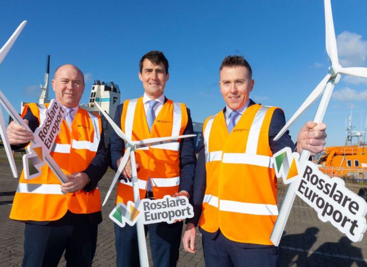 Three men standing outside holding plastic wind turbines and signs for Rosslare Europort.