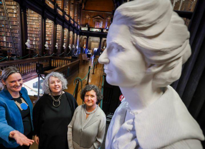 Three women looking at a sculpture of Rosalind Franklin in Trinity College Dublin's Old Library, with bookshelves in the background.