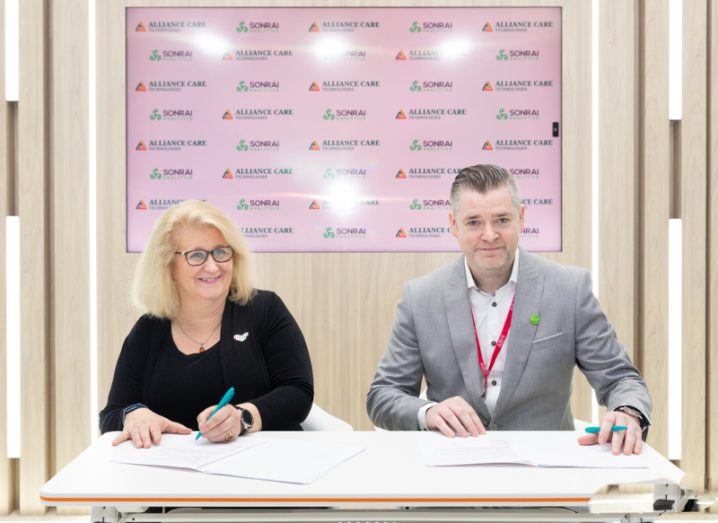 A woman and a man sit next to each other at a table and sign an agreement between ACT and Sonrai Analytics in the UAE.