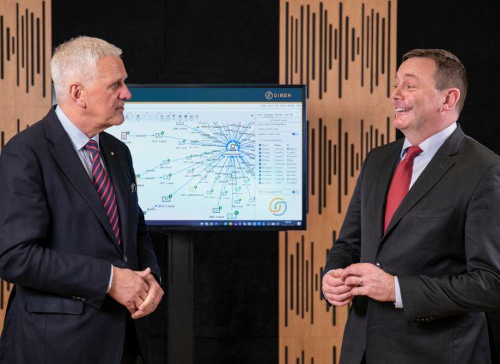 Two men wearing suits look at each other and smile. A monitor behind them shows the Siren platform on the screen.