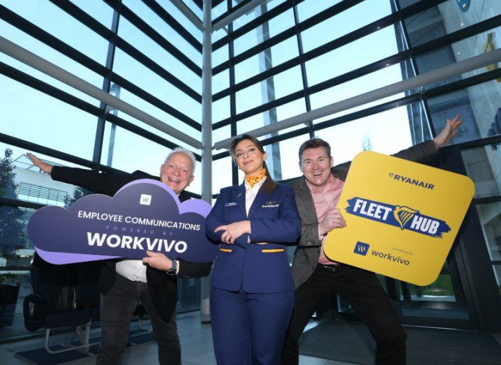 Ryanair DAC CEO Eddie Wilson and Workvivo CEO John Goulding hold up Ryanair and Workvivo branded signs and a woman in a Ryanair uniform stands in between them. There are large windows behind them.