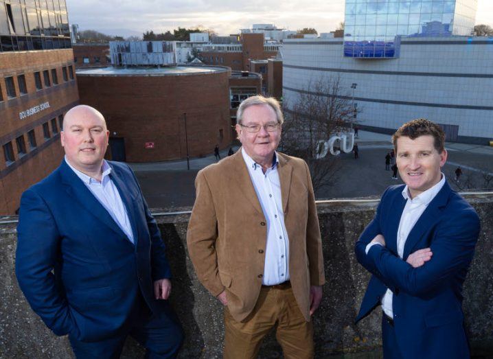 Three men wearing suits stand on the roof of a building with the DCU main campus visible in the background.