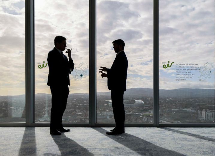 Silhouette of two men standing against the backdrop of glass walls inside a building with the Dublin skyline in the distance.