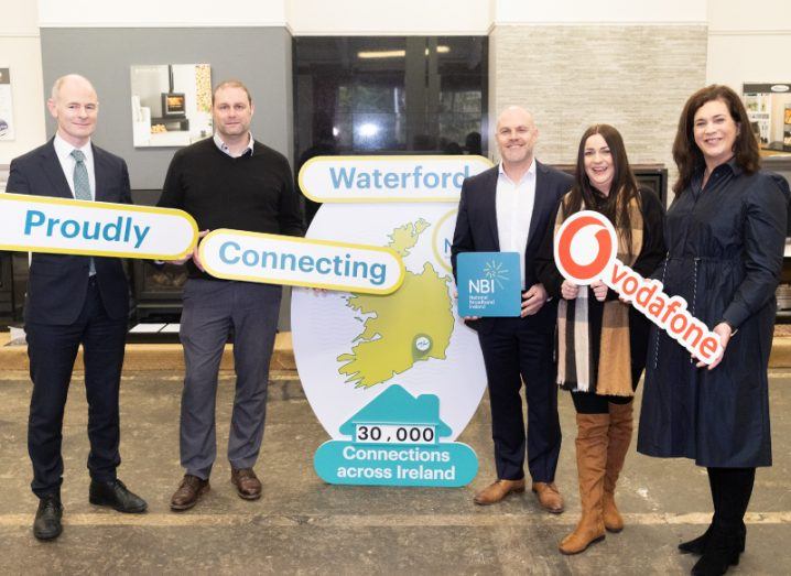 Three men and two women stand with a banner of Ireland in between them and holding signs to announce 'proudly connecting Waterford' with the NBI and Vodafone logos. There are in a fireplaces showroom.