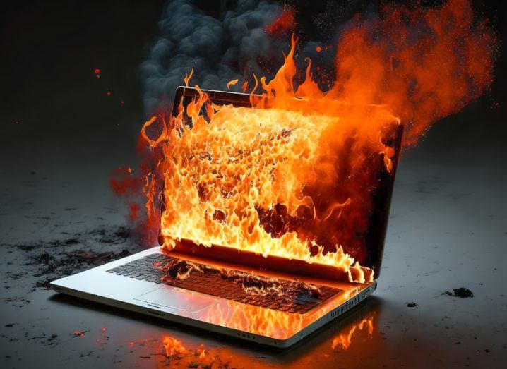 An open laptop that is on fire, with flames spilling out from the display and ash freckling the surrounding area.