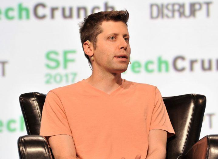 OpenAI CEO Sam Altman sitting on a chair with the TechCrunch logo on a wall in the background.
