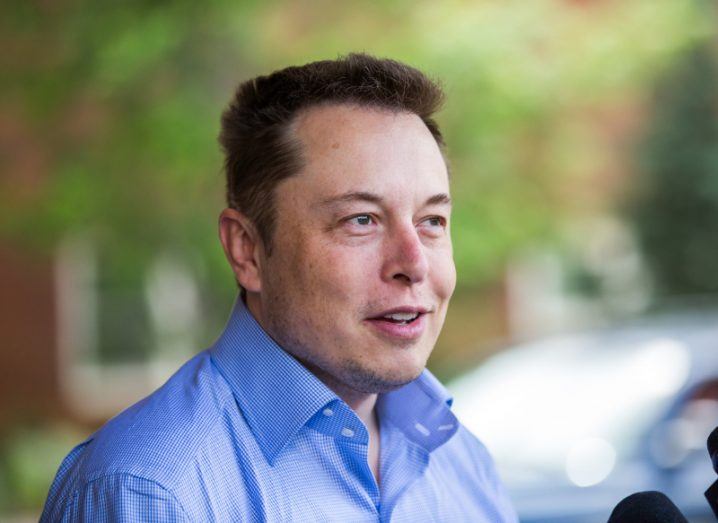 Elon Musk wearing a blue shirt, speaking in front of a small microphone with blurred trees and a car in the background.
