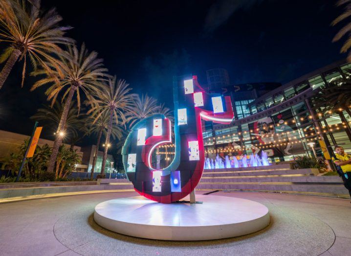 A statue of the TikTok logo, lit up on a circle stand with trees and a building in the background.