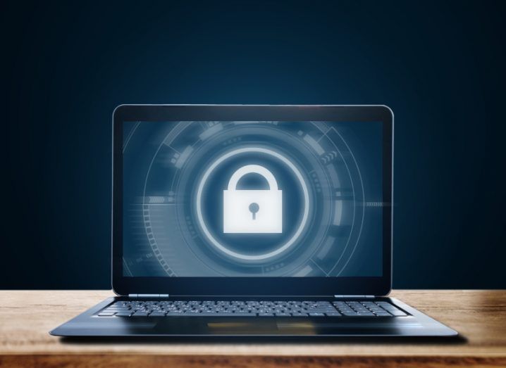 A laptop with a lock icon on the screen. The laptop is on a wooden table with a dark background behind it.