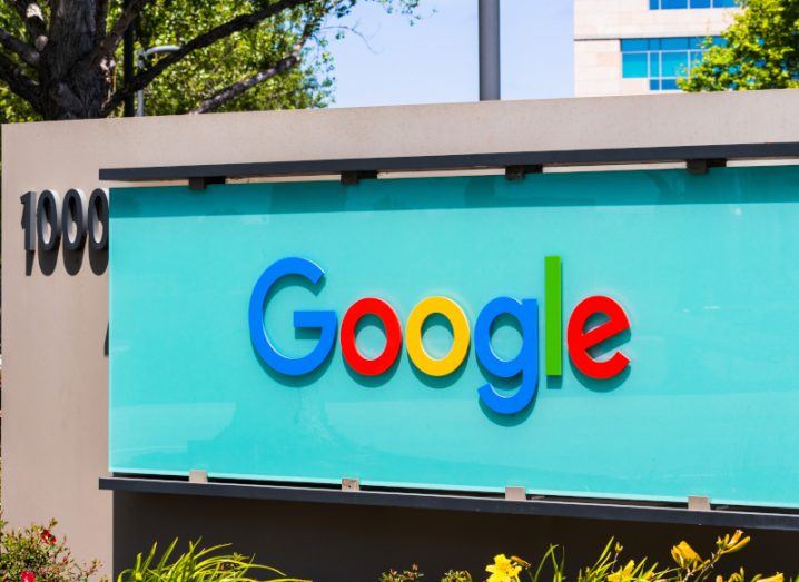 The Google logo on a blue sign, with trees, a building and a blue sky visible behind it.