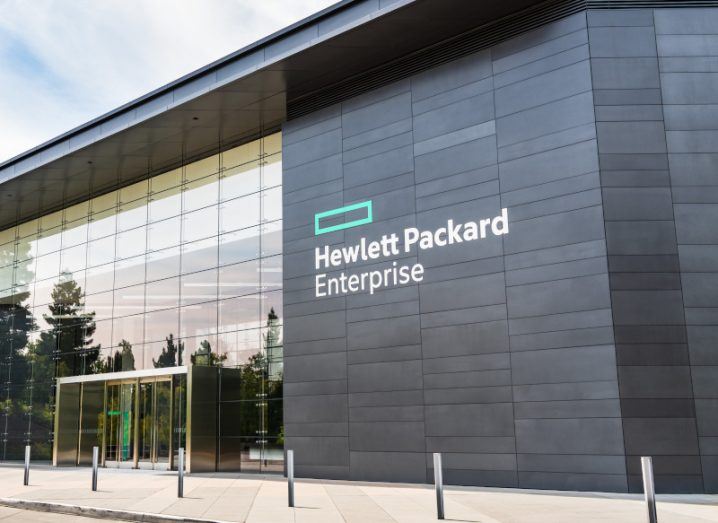 The Hewlett Packard Enterprise or HPE logo on the side of a black building, with trees reflecting off the building's windows and a cloudy sky above.