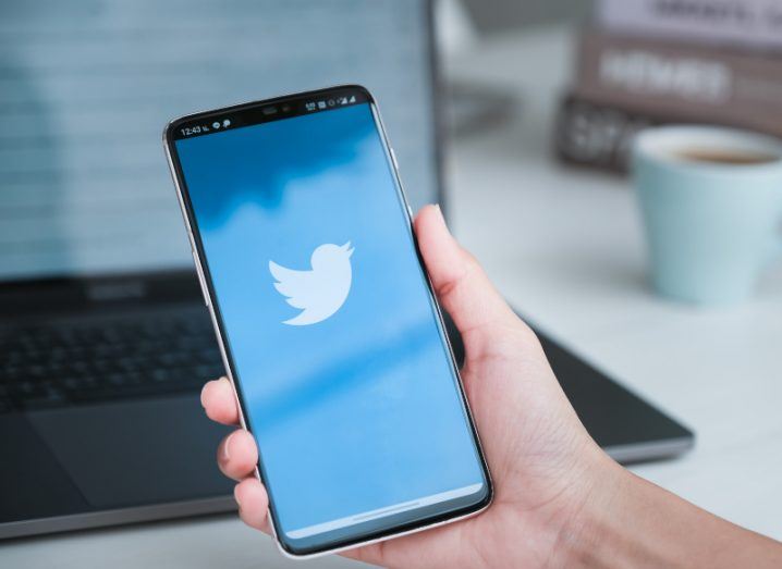 The Twitter logo on a mobile phone, held in a person's hand with a laptop and a cup in the background.