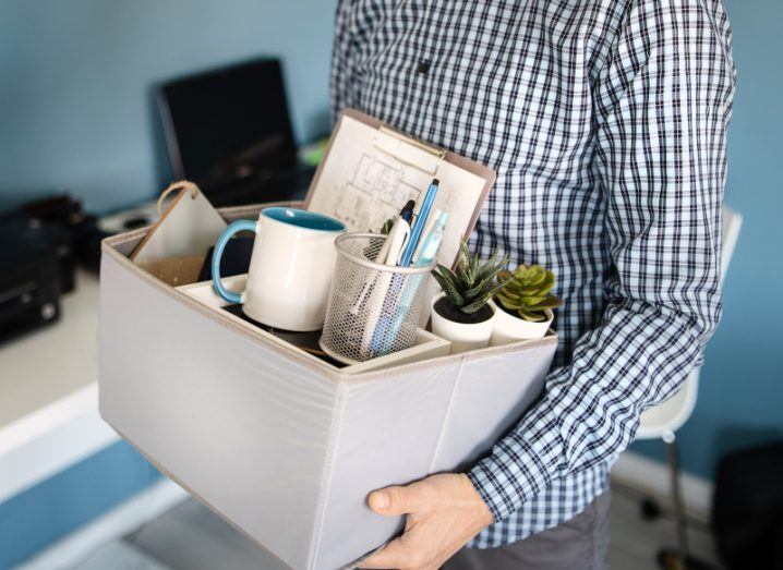 A person holding a box full of personal office belongings, such as a cup, pens and a notebook. Used as a concept of someone losing their job.