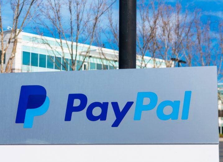 PayPal logo on a sign with a building, trees and a blue sky in the background.