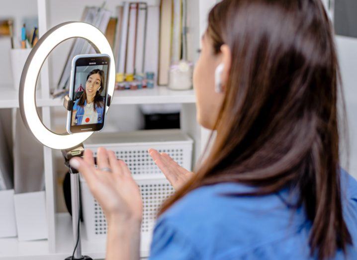 A woman recording a video on her phone, which is laying on a stand with a light behind it.