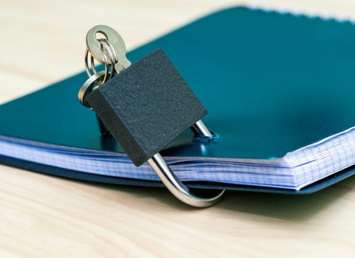 A closed notebook with a blue cover lying on a wooden table. A lock is attached to the notebook with a key inside it.