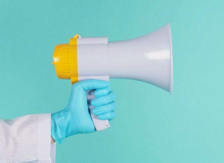 A megaphone being held in a person's hand in a blue background. The person is wearing a blue medical glove.