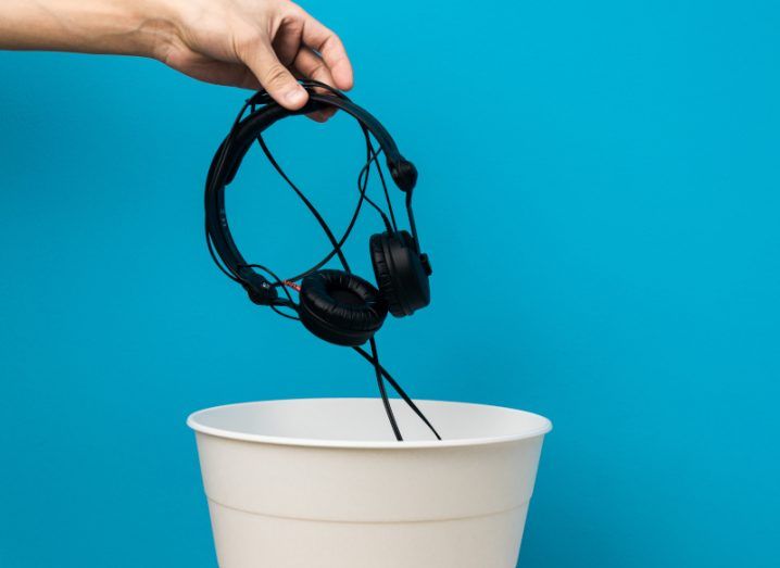 Person throwing a pair of headphones into a white wastepaper bin against a blue background.