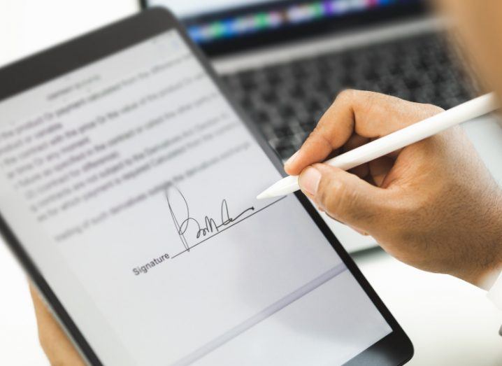 A person signing their name onto a digital tablet with a pen, with a laptop in the background.