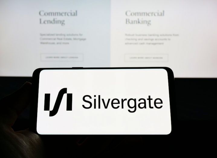 The Silvergate Bank logo on a smartphone screen, being held in front of a larger screen that shows the company's website.