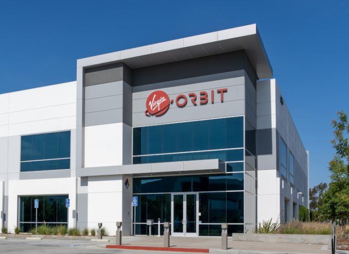 The Virgin Orbit logo above the entrance to a white building, with a clear blue sky in the background.