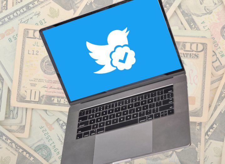 Laptop with Twitter logo and a verified checkmark on the screen with dollar bills in the background.