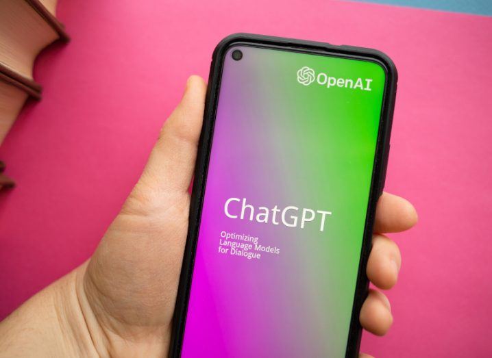 The ChatGPT and OpenAI logos on a smartphone screen, held in a person's hand with a pink surface in the background.