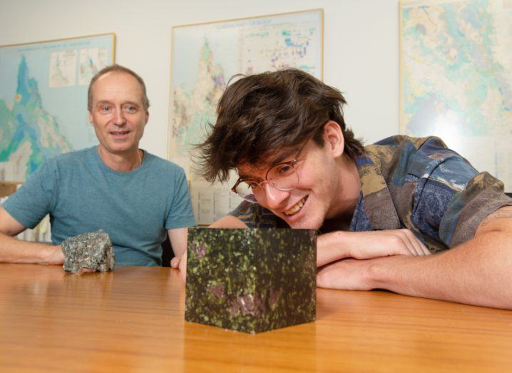 A researcher looking at a cube of rock on a table with another man behind him. There are maps on the walls of the room.