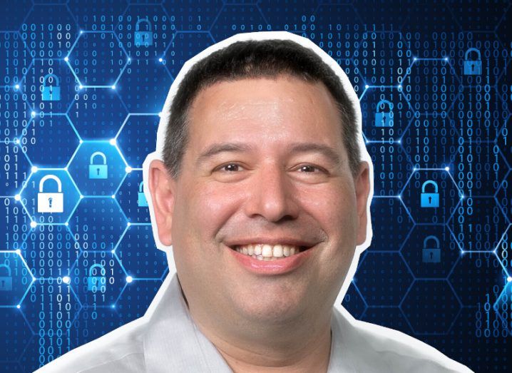 The headshot of a man smiling at the camera against a backdrop of blue padlock symbols surrounded by hexagon shapes.