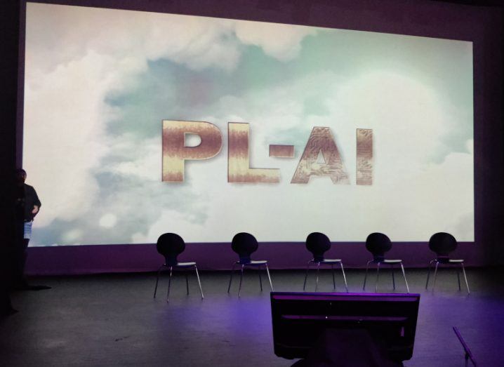 A screen showing the letters PL-AI, with a cloudy background. The screen is on the wall of a stage, behind five small chairs.